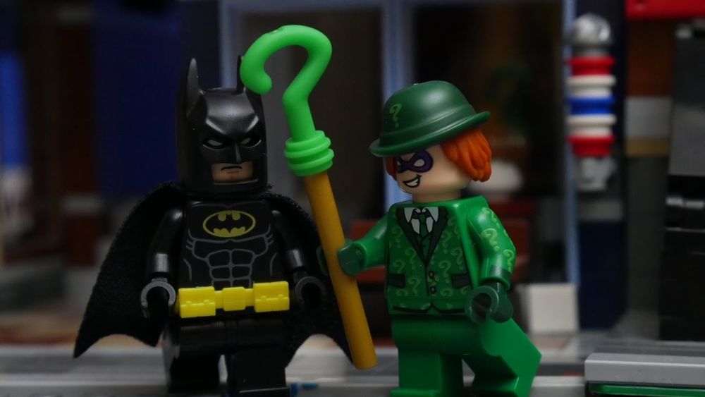 An image of the LEGO Riddler next to a LEGO Batman. He is seemingly tapping him with his question mark cane. The Riddler has an entirely green outfit, with lighter green question marks over his jacket and a single light green question mark on his hat. He has longer red hair and is grinning at Batman.