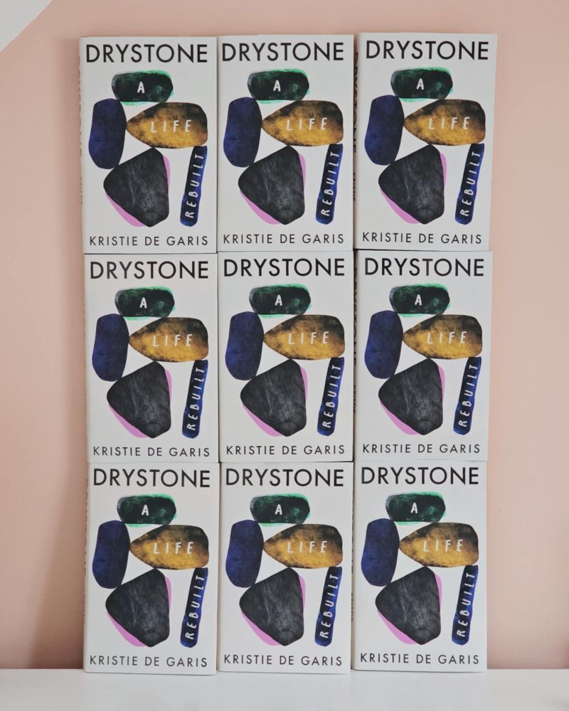 Nine front covers of the book Drystone – A Life Rebuilt by Kristie De Garis, arranged in a neat 3x3 grid against a soft pink and white background. The cover features abstract painted stones in black, green, gold, blue, and pink, with the title embedded across them.