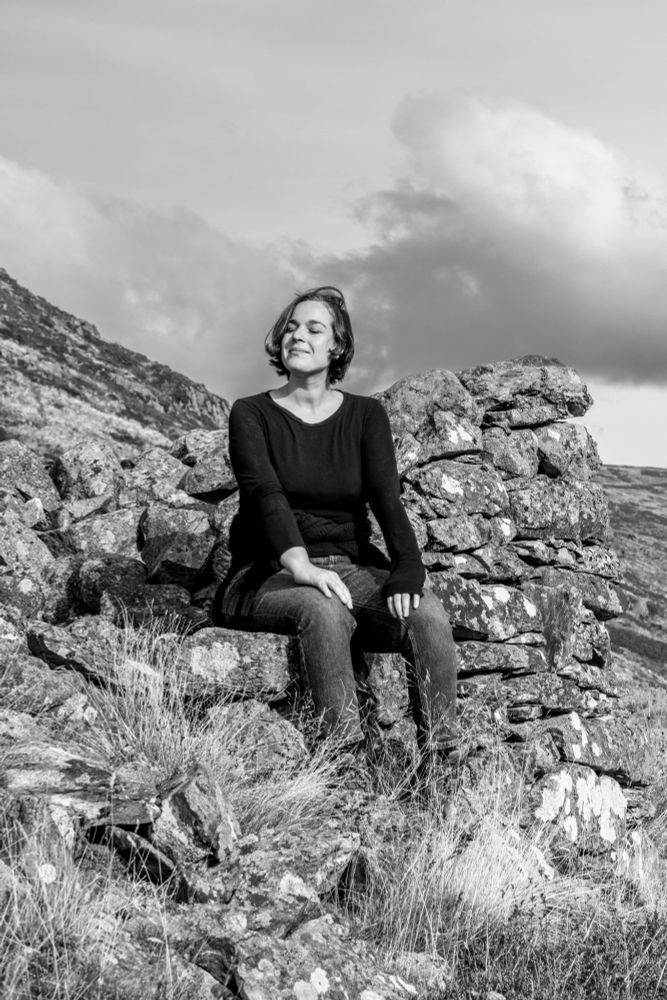Black and white photo of a woman sitting on a drystone wall in the hills, eyes closed and smiling gently. She wears a dark jumper and jeans, with wild grass and rough stone all around. The light is soft, and the mood is peaceful and quietly powerful. Photo by Christie Hemm Klok