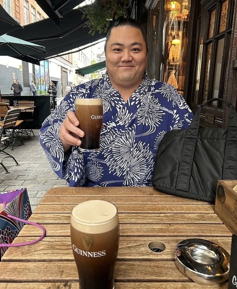 A sumo wrestler visiting London for a competitor in full traditional dress, sitting happily outside a pub with a pint of Guinness