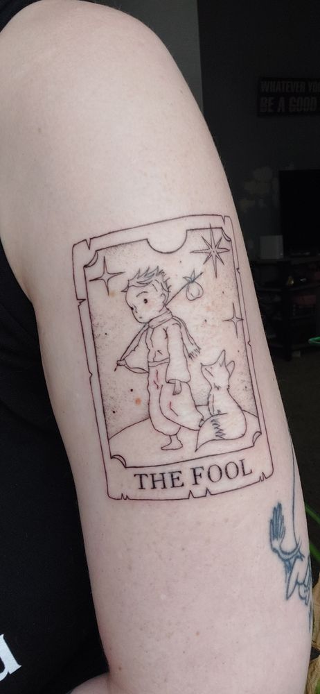 Tattoo of a tarot card on the arm. It shows the Little Prince as The Fool walking with a scarf and bundle, a fox at his side, and stars above.
