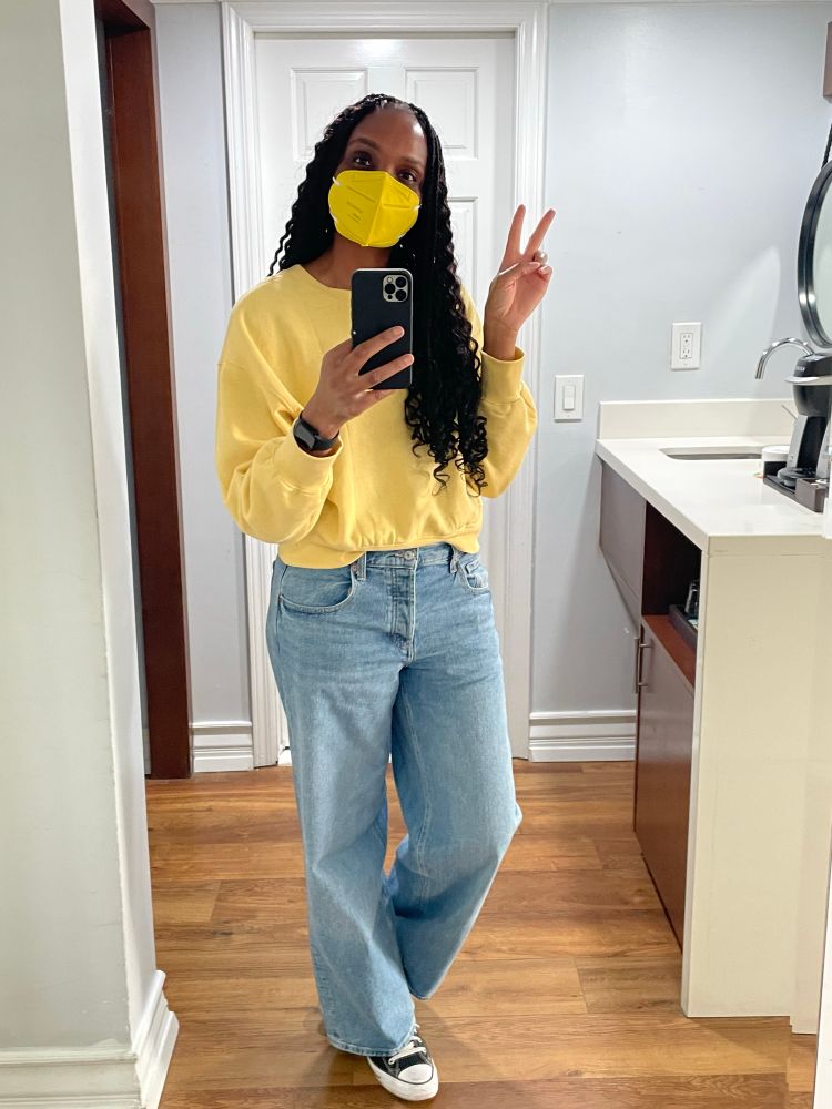 Tolu posing for a picture in front of a mirror, holding up a peace sign. She is wearing a cropped yellow sweater, baggy jeans, and a matching yellow N95 mask.