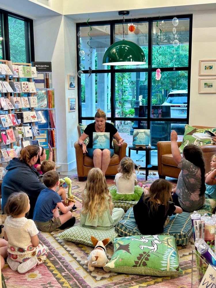Michelle sits at the front of a space facing a group of young children who sit facing her on a rug for story time. 
