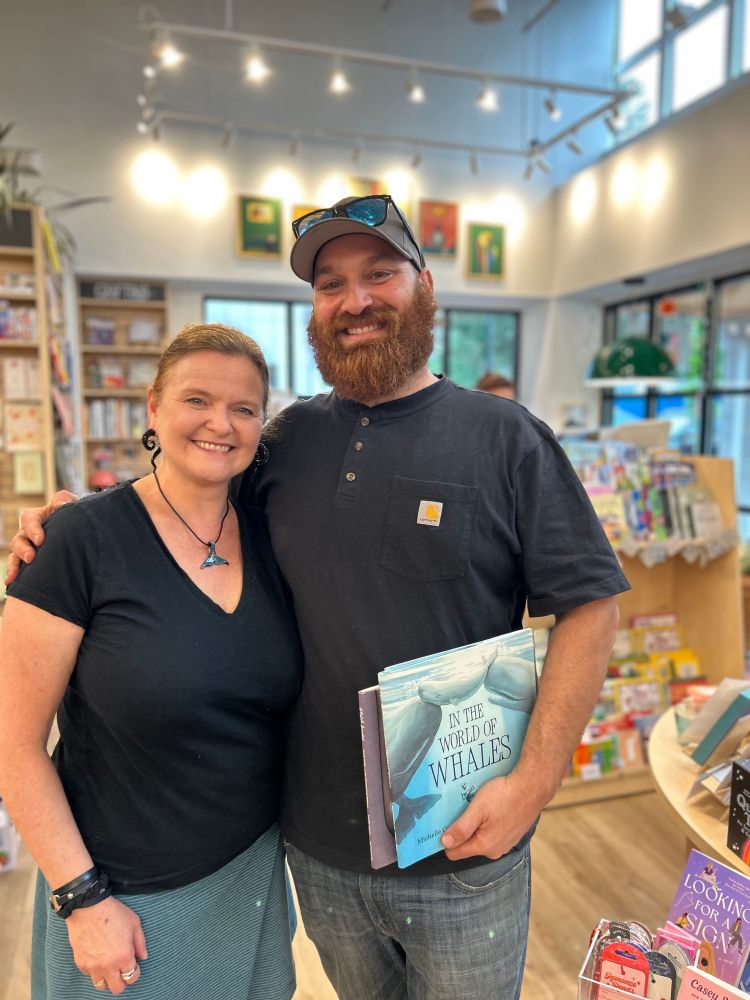 Michelle stands with her former student who holds a copy of IN THE WORLD OF WHALES. Both wear black shirts. Dan wears a ball cap and has a bushy reddish brown beard. 