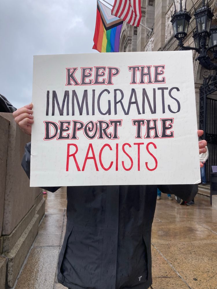 Person holds a sign that says “keep the immigrants. Deport the fascists.” A pride flag and a US flag are visible behind them. 