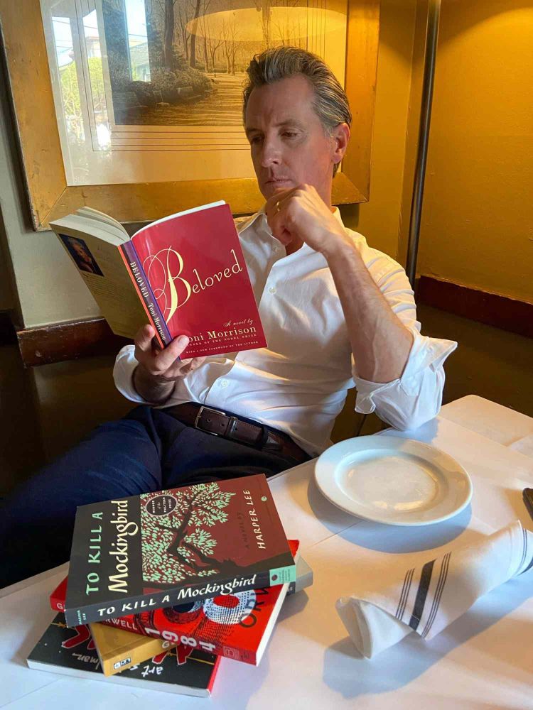 California Gov. Gavin Newsome, wearing blue pants and a white button-down shirt, pretends to be reading "Beloved" by Toni Morrison. He is seated at a table with an empty plate and a carefully stacked pile of four other books that he may pretend to read later.