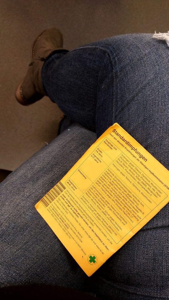 Man sieht meine Beine mit dem Impfpass im Schoß 