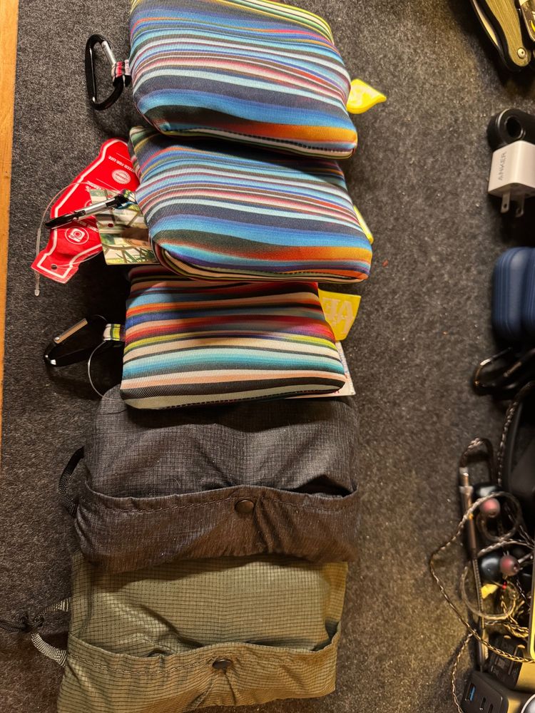 A picture of five portable tote bags folded up on a cluttered desk. Three squarish tote bags are colorful stripes, while the other two are gray and olive. 