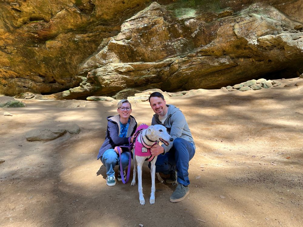 Photo of Bree and Alex, two people with white skin and short hair, crouched down smiling and posing with Kona a white greyhound. The ground is sandy and behind them is a wall of sandstone. At Ash Cave at Hocking Hills State Park. 