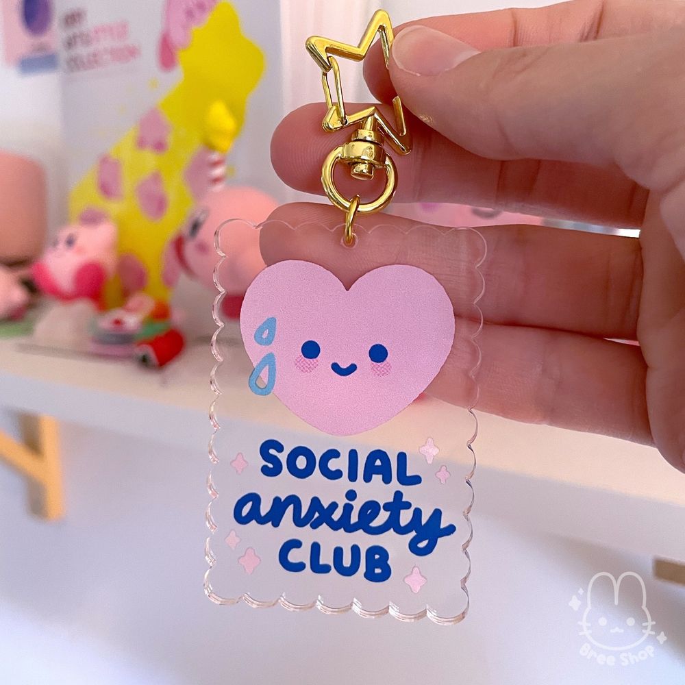 Photo of a hand holding up a rectangular clear acrylic charm with scalloped edges and a cute pink heart on it. The heart is smiling and sweating and below it says “social anxiety club” in blue hand lettering. It has a gold clasp in the shape of a star.