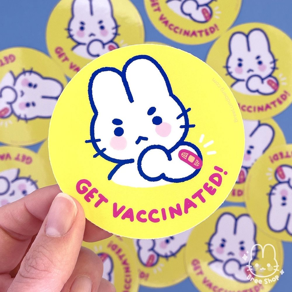 Photo of a hand holing a round bright yellow sticker with a cute illustrated white bunny character looking determined showing off their arm with a bandaid on it. Below the bunny it says "get vaccinated!" in bold pink lettering.