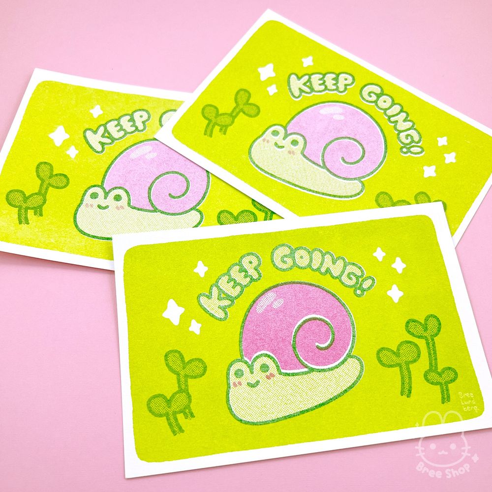 Photo of risograph prints of a cute illustrated pink snail on a lime green background. The snail looks happy with sprouts around it and bubble letters that say "keep going!" above it.