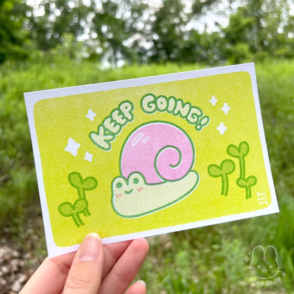 Photo of a hand holding up a small risograph print of a cute illustrated pink snail on a lime green background. The snail looks happy with sprouts around it and bubble letters that say "keep going!" above it.