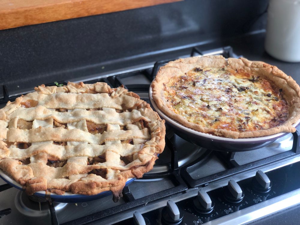 Lattice top apple pie with an uneven lattice and wonky crust edge beside a more respectable quiche. Both in stoneware pie dishes, sitting on a stove.