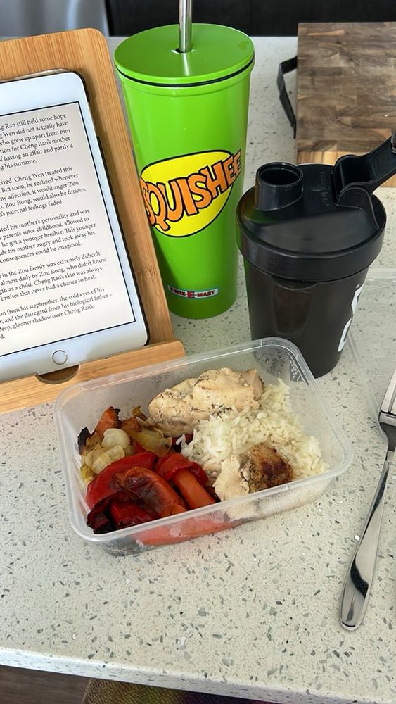 Sad baked chicken with rice and roast veges in a plastic container with shake and metal cup in background. To left is iPad displaying some midrange nugu danmei.
