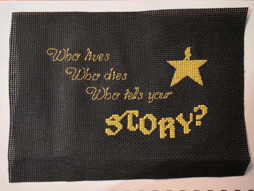 Who lives Who dies Who tells your done in golden backstitch, STORY? done in cross stitch in the same color. A golden "star" with a small figure atop it representing the Hamilton musical logo. All done on black 14-count aida fabric. 