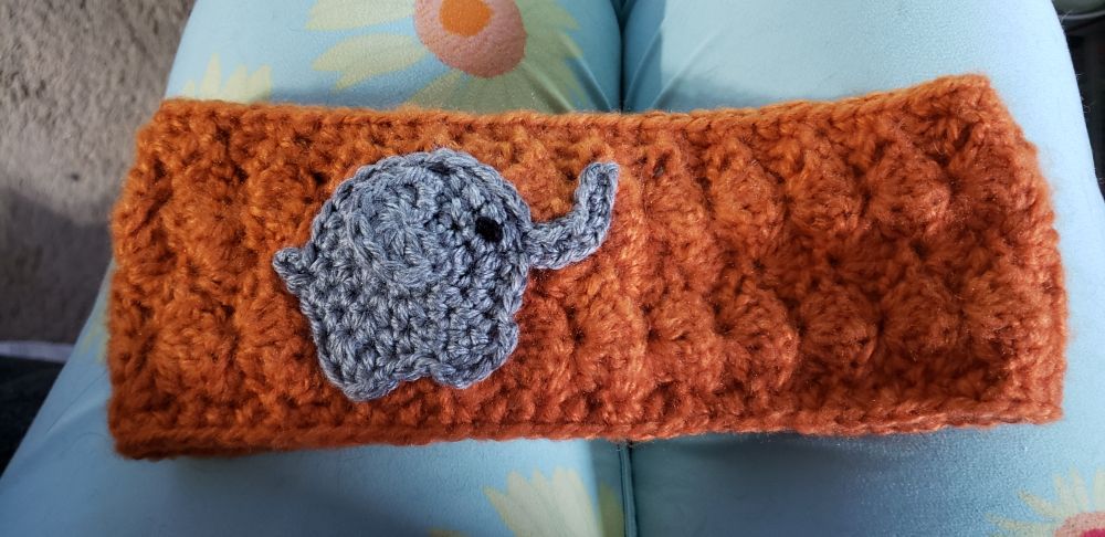 A grey, crocheted elephant applique on an orange ear warmer headband done in a shell stitch.
