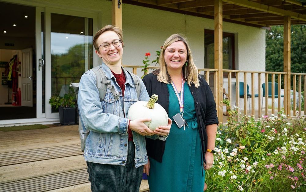 Two women laugh and pose for the camera while holding a large blue squash next to a wildflower bed
