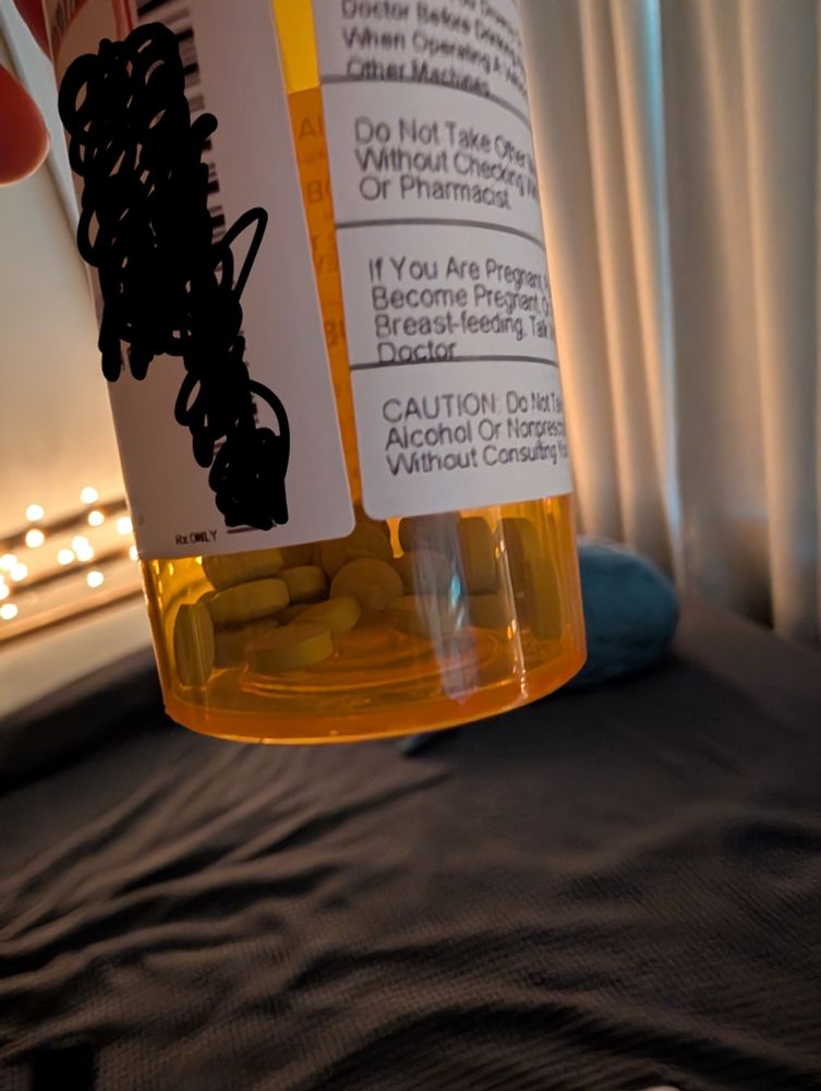 An orange bottle with small blue pills. It's held in front of a cozy grey bed with curtains, string lights, and a blahaj