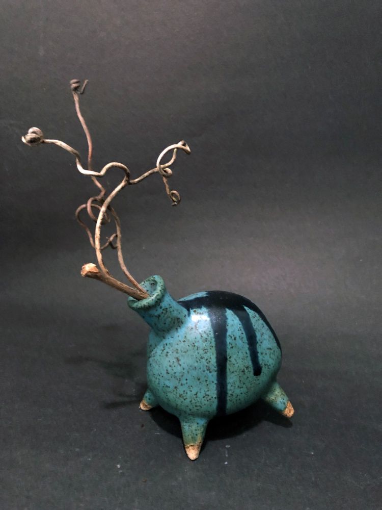 Small turquoise ceramic bottle with black drips from back running down the sides. Bottle is poised on 4 little peg legs with one back leg raised as if in motion. Small neck is placed off center, further giving an animalistic appearance, and a few curly twigs from wild grape vines emerge from the neck.
