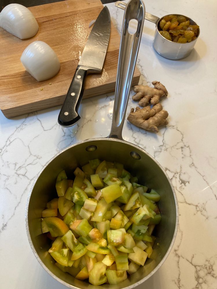 Overhead view of a kitchen counter with a pot of chopped green tomatoes and a cutting board with a white onion cut in half and a chef’s knife. There is also some ginger root and a cup of golden raisins.