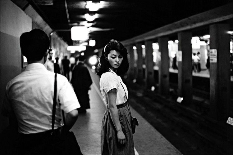 Black and white photograph of a Japanese woman, wearing a white blouse, dark skirt, stands on a subway platform, sideways on, looking slightly downwards, towards the camera. There are other travelers also walking around on the platform. The train is probably approaching as you can see a distant light source. It's a very contrasty image, rich in blacks. Image taken by Ed Van der Elsken, titled Girl in the Subway, Tokyo 1981