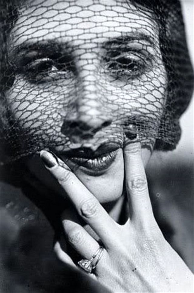 Black and white photograph of an attractive woman, wearing a veil, her hand and fingers are in a V position in front of her face. Image by  Jacques-Henri Lartigue (1894-1986).