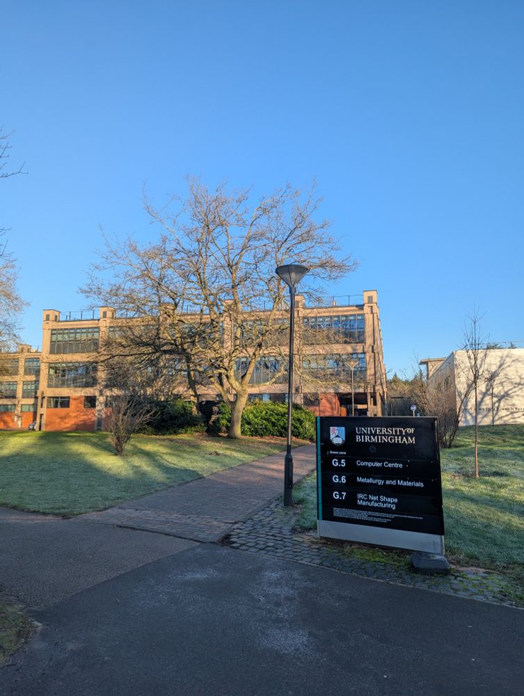 Picture of the School of Metallurgy and Materials at the University of Birmingham. A brown building with a tree in front and a sign, bright blue sky.