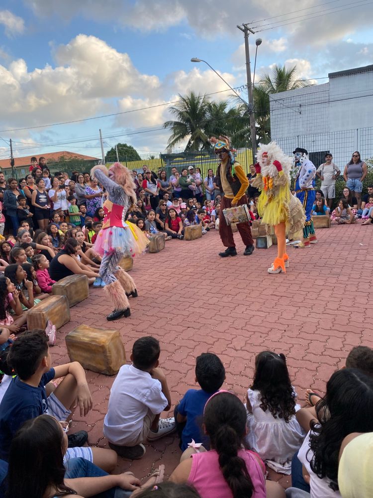 Uma roda de crianças em volta de atores vestidos de gato, jumento, galinha e cachorro.