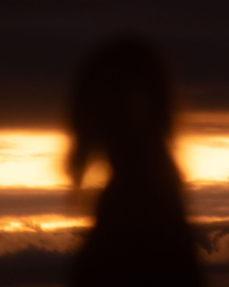 A digitally created blurry silhouette of a girl standing sideways against a sunset backdrop. The deep black silhouette contrasts against vivid and richly orange glowing clouds and sky. The hazy and indistinct forms blend softly, expressive of abstract photography and digital art. Colors shift between dark black, deep browns, and warm golds, generating a moody and atmospheric sense of memory or wistfulness.