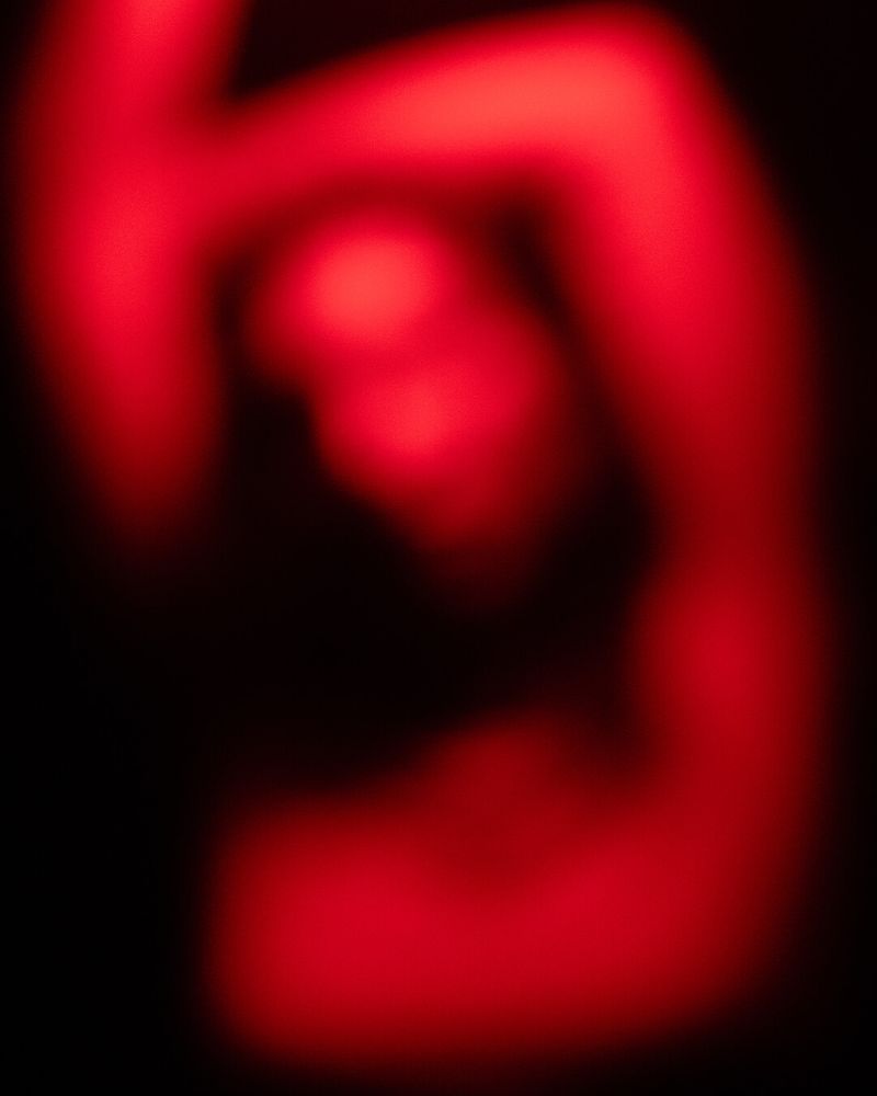 Abstract photography depicting an out-of-focus human figure colored in vivid red lighting against a stark black background. The silhouette is indistinct and blurred, showcasing raised arms and suggesting a dynamic pose. The composition concentrates on intense color contrasts of deep blacks and radiant reds, evoking mystery. Image utilizes techniques in experimental digital art and abstract photography.
