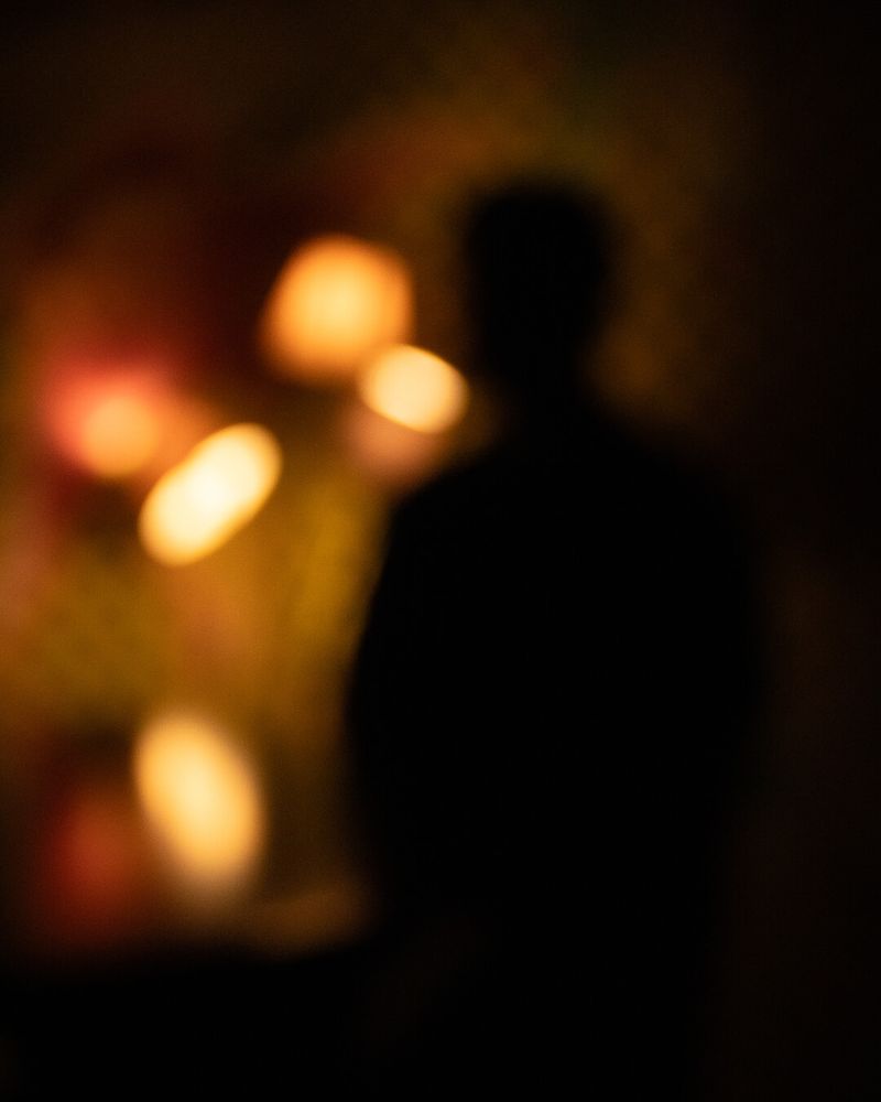 An abstract, blurry photograph showcasing the dark silhouette of a figure against a softly illuminated background with warm, golden hues. Diffuse circles of blurred yellow and orange lights softly glow, creating a dreamy, unclear ambiance. The composition conveys mystery through its intentional lack of focus, leaving details to imagination. Artistic digital photography capturing silhouettes and abstract lighting effects.