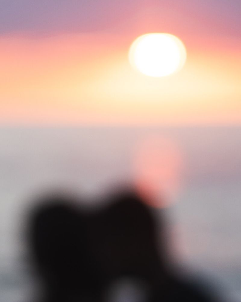 Abstract, out-of-focus photography of two silhouetted figures embracing against a backdrop of a blurred pastel sunset. Warm gradient hues of pinks, oranges, purples and yellows blend softly, emphasizing emotional intimacy and calmness. There is heavy blur typical of digital art that resembles a dreamy, memory-like quality, creating a gentle, romantic atmosphere.
