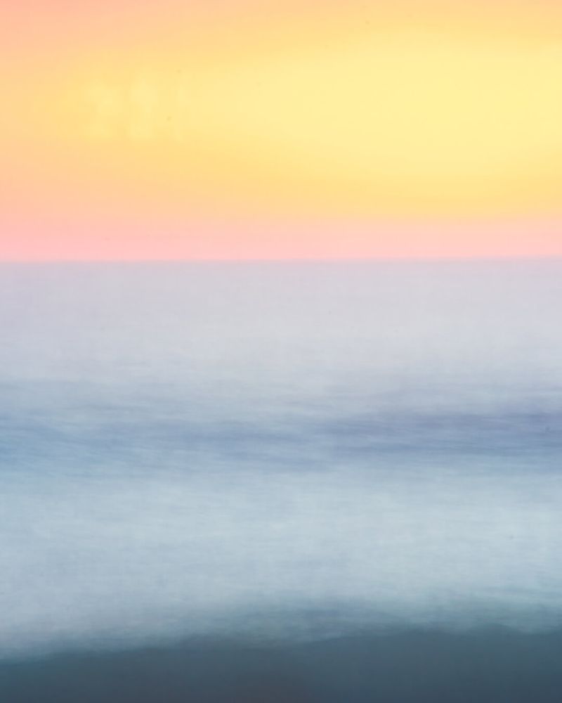 Abstract, softly blurred digital photograph depicting a sunset at the beach. The image is layered horizontally with gentle gradients of warm yellow and peach blending towards soft pink above, transitioning into muted shades of blue and turquoise representing the sea. The image evokes a dreamy and calm atmosphere, showcasing an experimental photography style focused on long exposure and blurred clarity. Ideal for digital art enthusiasts.