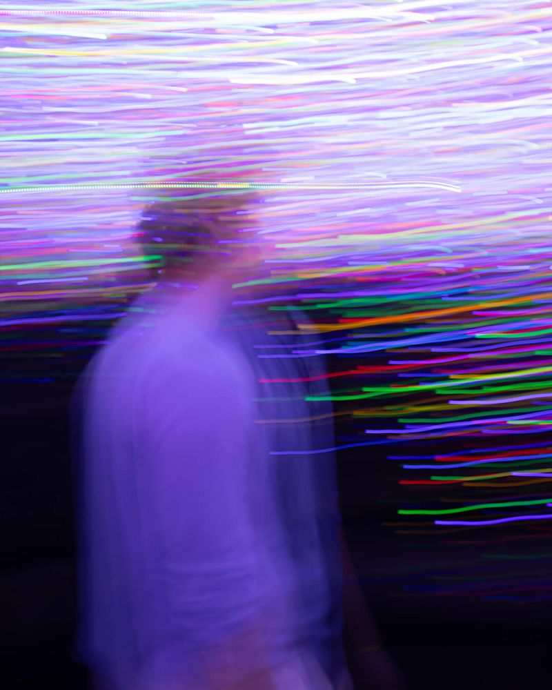 A blurred image with motion blur of colorful neon lights streaking horizontally across the frame. In the foreground, a person, partially visible, is indistinctly superimposed against the background, adding depth. The colors primarily involve vibrant shades of purple, pink, blue, and hints of green. The lower portion transitions into deeper shadows, providing contrast. Technique indicative of abstract digital art photography, evoking an experimental visual style.