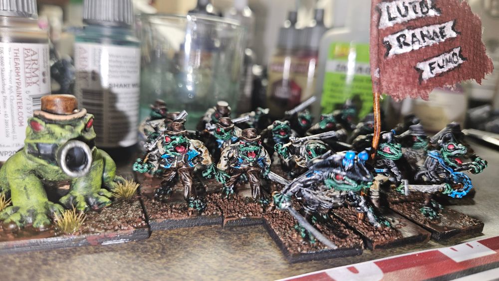 Painted miniature anthropomorphic frogs in British naval uniforms carrying shotguns, one is very large with a cannon in its mouth. A standard bearer carries a flag with the motto 'LUTO RANAE FUMO' which I think translates to 'MUD FROGS SMOKE'