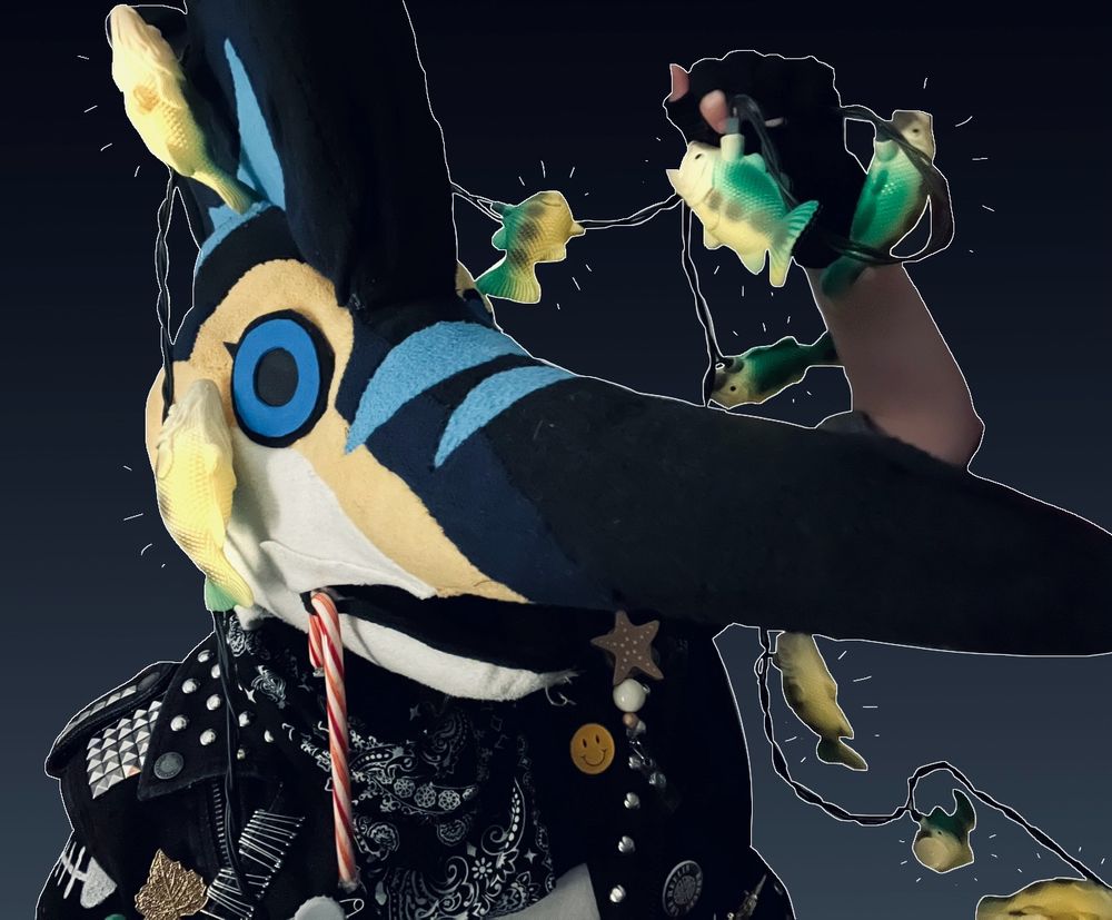 A photo of myself with my blue marlin fursuit had on, tangled up in Christmas lights that are shaped like fish, and wearing a battle jacket. The background is edited to be a black to grey gradient. There is a candy cane hooked in my fursuit head’s lip.