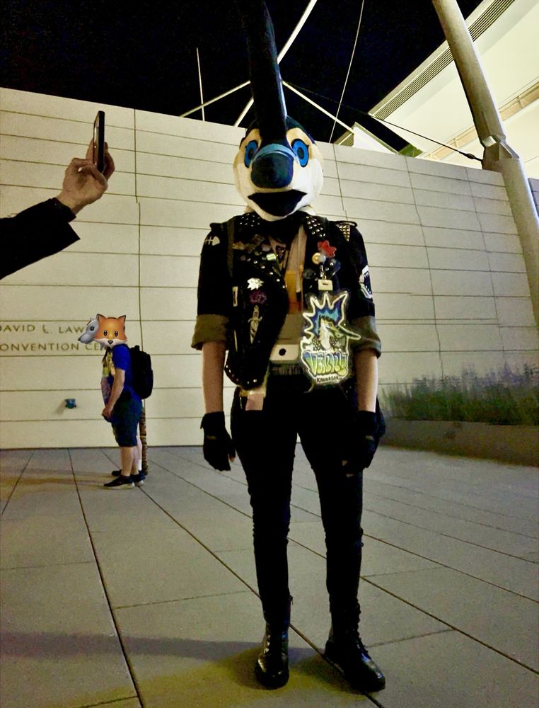 My standing facing the camera. I’m wearing my Marlin fursuit head, battle jacket, skinny jeans and black boots. Someone is holding a phone camera close to my head and there are two bystanders in the background. 