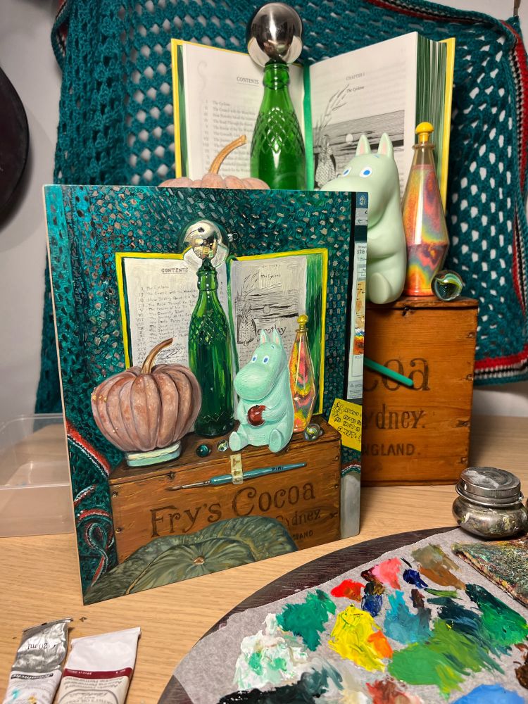 Oil painting featuring a futsu pumpkin, green bottle, mirrored bauble, moomin toy holding an Apple, a coloured sand bottle, and marbles, all on top of a wooden Fry’s Cocoa box and behind these items is an open yellow book (The Wizard of Oz) with green page edges. There is a green crocheted blanket with red, white and black trim as the back drop. In front of the Cocoa box is a green striped pumpkin. The painting is on a desk with a full palette and the painting set up behind it.