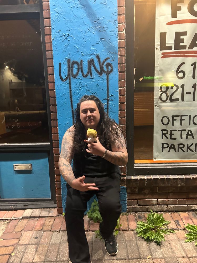 Person in black tank top and pants crouching in front of graffiti that says YOUNG they’re holding a ice cream in their hands and there are two panes of glass of the shop they’re standing in front of 
