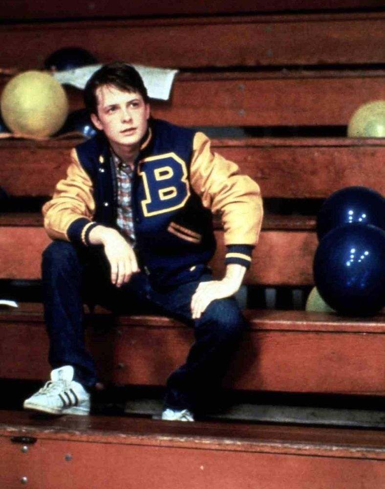 A man sits on the beaches of a school gym. He wears a blue and yellow letterman jacket with the letter B on it over a battered shirt. His jeans are dark blue, and his shoes are white with black details.