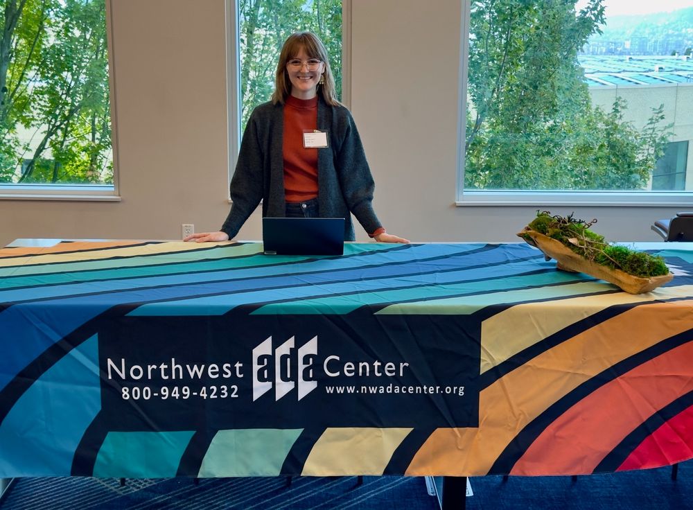 Kaitlin standing behind table with rainbow patterned Northwest ADA Center vendor table.