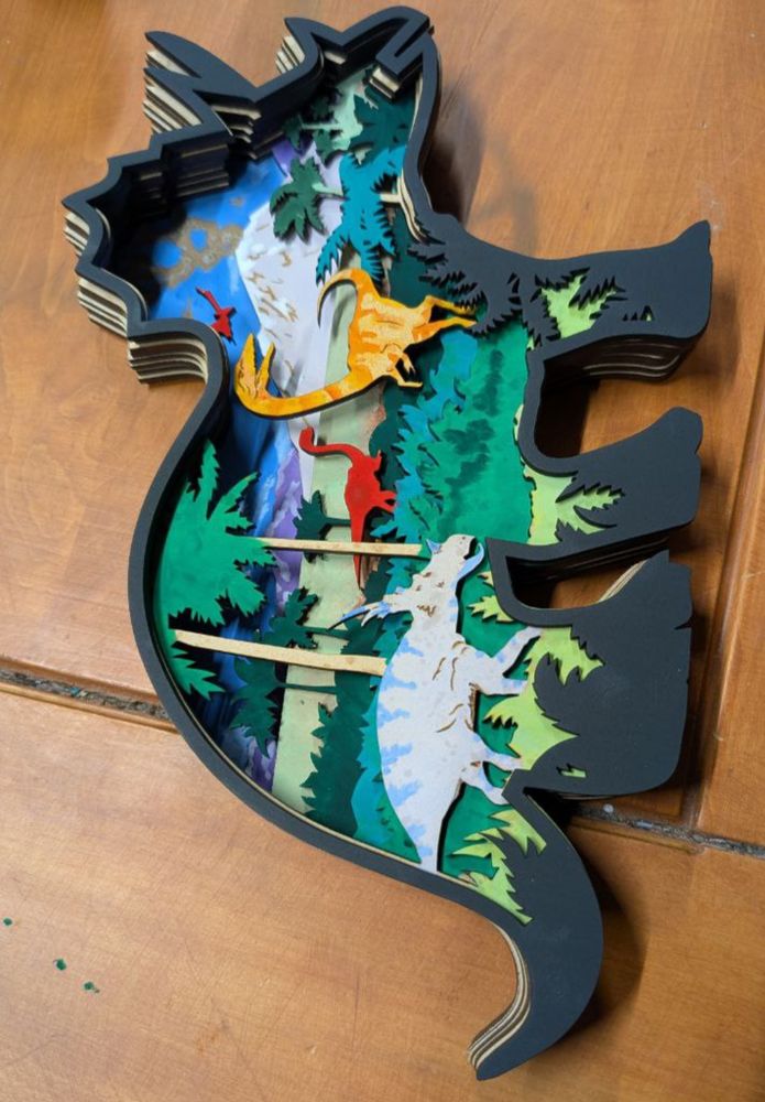 A wall art decor piece in the shape of a triceratops silhouette made of nine layers of MDF cut out with a laser cutter. These layers are glued so the designs over lap and make up an image. The image is of a volcano smoking on the horizon with several dinosaurs and Jurassic like plants closer to the viewer.