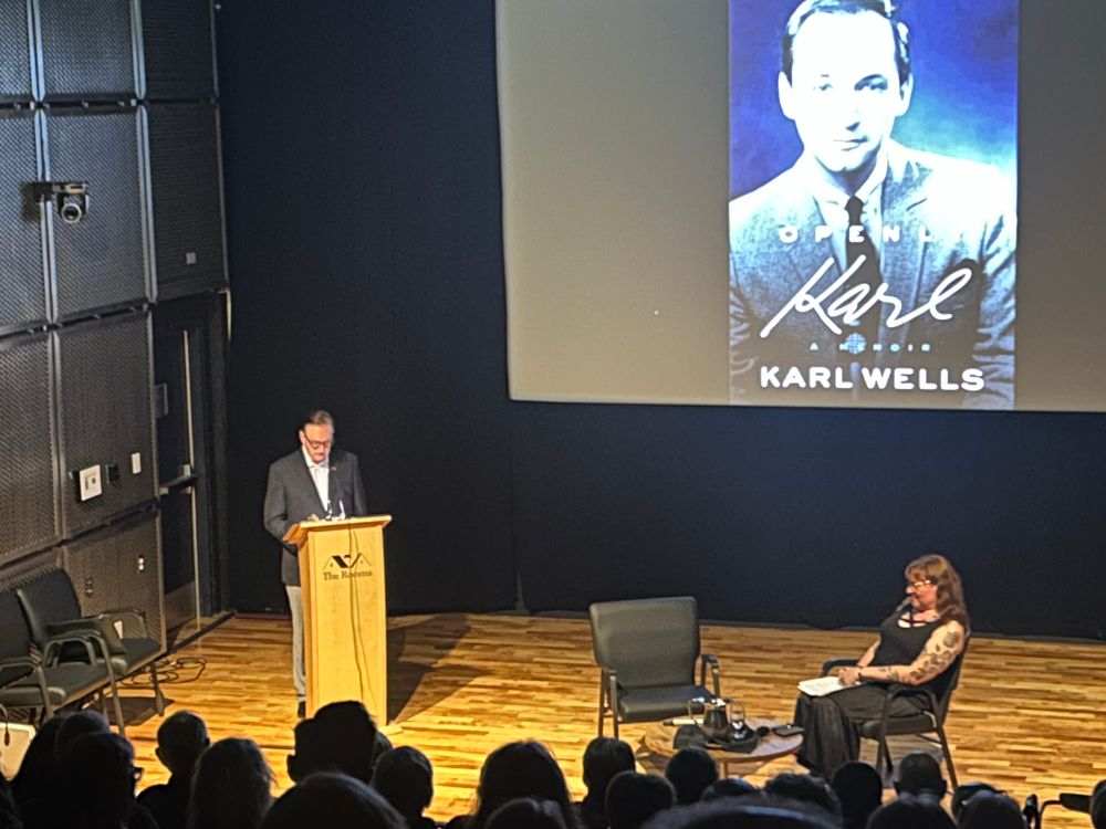 Karl Wells speaking at a podium, with Rollmann sitting, and Karl’s book cover, Openly Karl, projected in the background