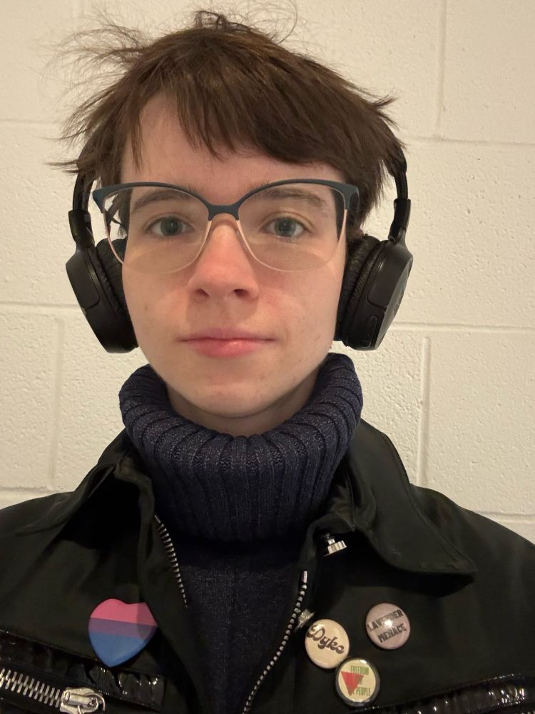 Selfie of me (nora) in a dark blue turtleneck and my bi jacket. I’m wearing my headphones and the background is cinderblock