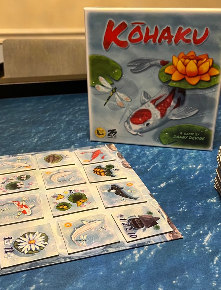 The box for Kōhaku featuring a white and red koi fish swimming by a yellow flower on a Lilly pad and a dragonfly nearby. In front of the box is a grid of cardboard tiles alternating koi of various colors and different koi pond features such as turtles, flowers and butterflies. 