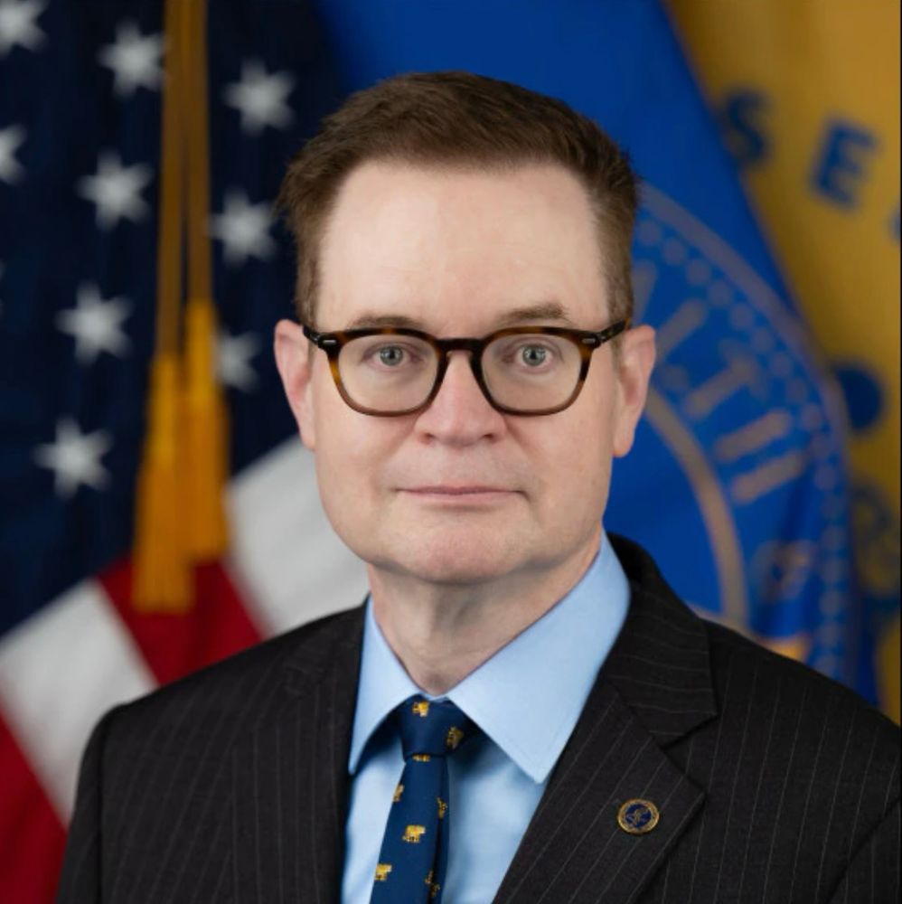 Photo of a white mab with short reddish hair who is wearing a charcoal pinstripe suit. He is posed in front of the American flag and is wearing squarish tortoiseshell glasses 