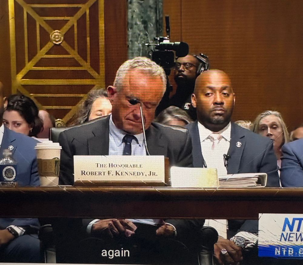 Kennedy in a grey suit sits behind a name card and is texting on his phone under the table . 