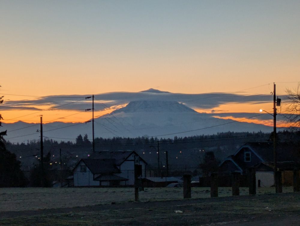 A snow-covered mountain with a line of clouds across its top. The sky around it is slowly turning orange as the sun rises.