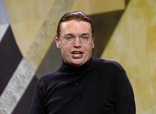 A screenshot of comedian Mike Myers in his role as Dieter in the recurring Saturday Night Live sketch SPROCKETS, the self-same fictional West German talk show of which Dieter is the host. Dieter is wearing a black turtleneck and wireframe glasses and sports slicked-back hair. His mouth is open as if he's mid-sentence, implying that he's stating the quoted post in his trademark flat German accent. He stands in front of an abstract black, gray, and yellow shapes on a mural, painted in what is likely an expressionist or Bauhaus style.